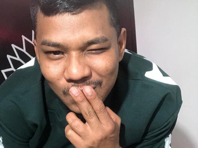 SamSexy69 mit kurzen, schwarzen Haaren, einem leichten Augenzwinkern und einem kleinen Schnurrbart, der sanft an seinem Finger knabbert, trägt ein grünes Sportshirt und zeigt einen verspielten, neugierigen Ausdruck..