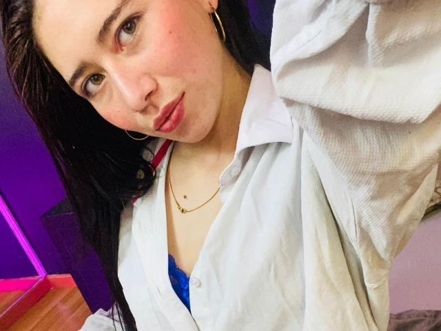 LexyJones, con piel pálida, cabello oscuro y aros grandes, vestida con una camisa blanca que deja ver un sujetador azul, labios fruncidos y un collar dorado, mirando a la cámara..