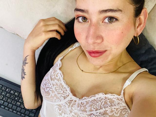 LexyJones, con pecas, ojos marrones y cabello negro largo, vestida con lencería blanca de encaje, aros de oro y un collar, acostada en un sofá con una computadora portátil. Se ven los senos..