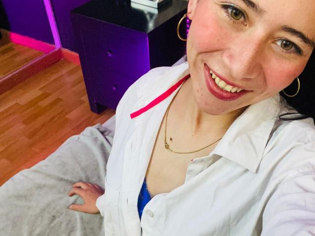 LexyJones with a smile, wearing a white shirt over a blue bra, showing her tits, hoop earrings, and a necklace, sitting on a gray blanket, wooden floor.