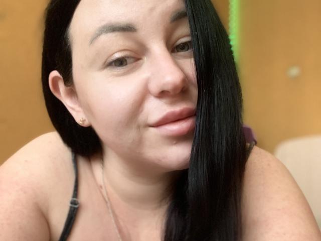 VeronikX hat eine blasse Haut, schwarzes, glattes Haar, das ein Auge bedeckt, kleine Ohrringe und ein kaum merkliches Lächeln mit leicht geöffneten, rosafarbenen Lippen. Sie trägt ein schwarzes Top mit Trägern..