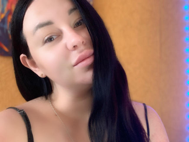 VeronikX a les cheveux noirs, une peau claire, de petites boucles d'oreilles, et elle regarde légèrement sur le côté avec un sourire discret. Elle porte un haut noir à bretelles fines..