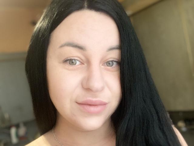 VeronikX heeft lang zwart haar, een lichte huid, groene ogen en roze lippen. Ze draagt een zilveren ketting en heeft een subtiele, kalme uitdrukking op de foto..