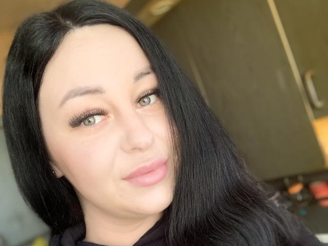 VeronikX ha lunghi capelli neri, occhi verdi e rossetto rosa. Guarda di lato con un leggero sorriso, indossando una maglietta nera, che mette in risalto il suo viso carino..