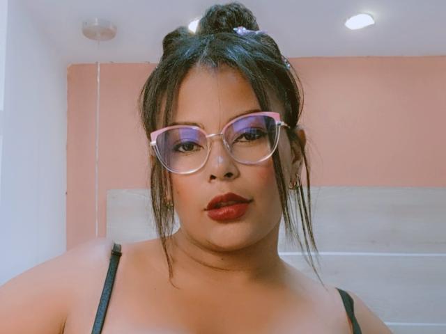 Mikella, con occhiali rosa, rossetto rosso, reggiseno nero e capelli castani raccolti in uno chignon, guarda verso la fotocamera, mostrando il suo viso paffuto e i suoi piccoli seni..
