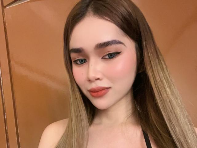 GabbyHottieVixen with long brown hair, blue eyes, winged eyeliner, pink blush, and orange lipstick, looking down with a slight smile, showing her small, cute, and slightly parted lips.