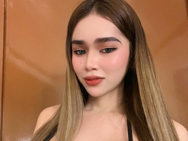 GabbyHottieVixen has long brown hair, thick eyebrows, black eyeliner, pink blush, red lipstick, and a small mole on her left cheek, wearing a black top, looking slightly down.