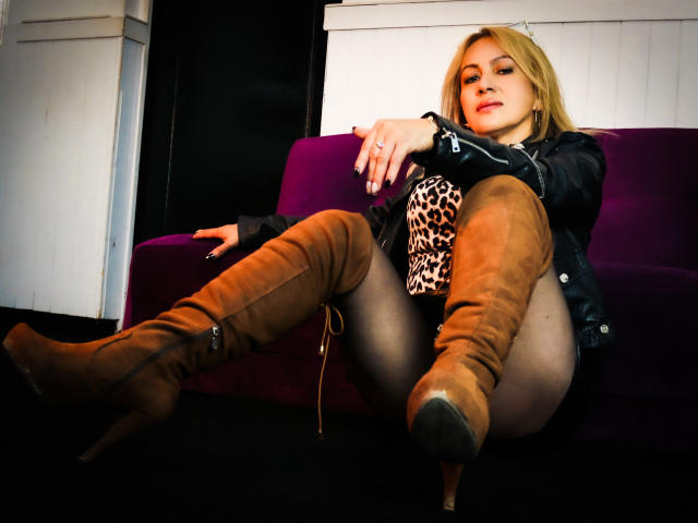 LindaWalton with blonde hair, wearing a black leather jacket, leopard print dress, and brown thigh-high boots, legs spread, sitting on a purple couch, looking seductively at the camera.
