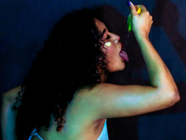 SophieSonyas with curly black hair, tongue out, holding a yellow-green banana to her mouth, purple nail, white face paint, wearing a blue top, dark background.