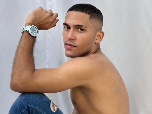 DereckStorm is shirtless, with a toned, brown-skinned, muscular upper body, short black hair, and a light blue watch on his right wrist, sitting and looking at the camera.