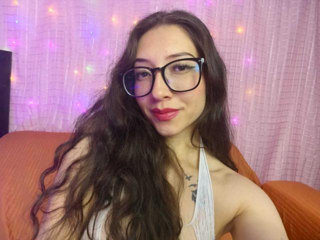 ElizabethMorrison with long wavy brown hair, wearing black glasses and a white halter top, showing her tits and a small tattoo on her chest, smiling with pink lipstick.