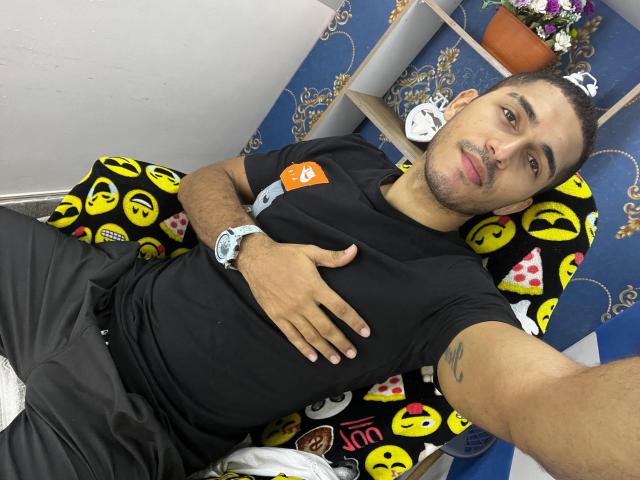 DereckStorm met kort donker haar, een zwart T-shirt, zwarte broek en een horloge, liggend op een geel emoji-kussen, met een hand op zijn buik, een tatoeage op zijn rechterarm, en kijkend naar de camera..