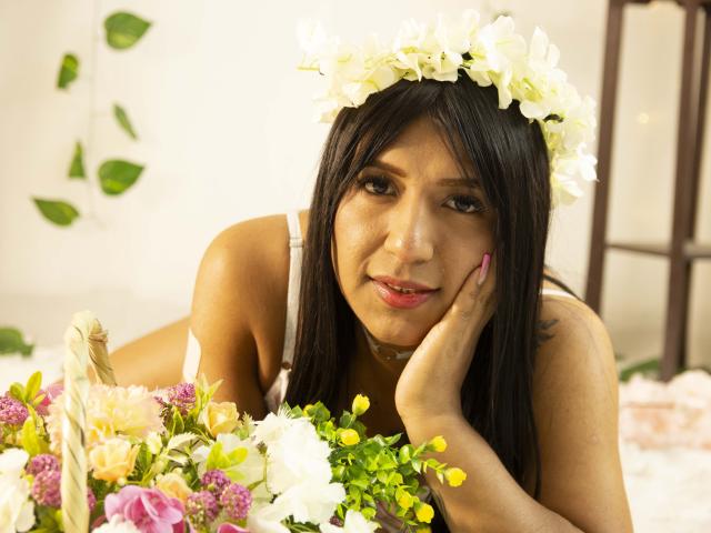 IsabellaStonnee with a white flower crown, lying down, head resting on hand, looking at camera, pink nails, brown eyes, black hair, white dress, bouquet of flowers in front.
