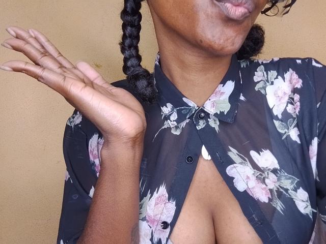 MariaTomas, met donkere huid, gevlochten haar, draagt een zwart bloesje, haar hand is omhoog, ze laat haar decolleté zien, het shirt is gedeeltelijk open en haar borsten zijn zichtbaar. Beige achtergrond..