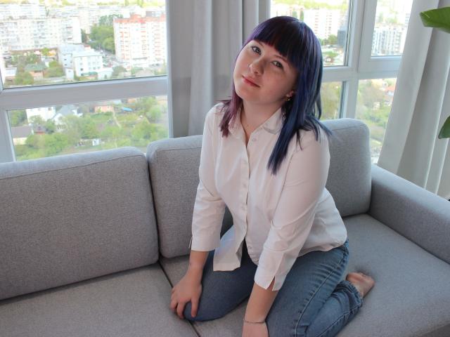 Felizia4U-hot with purple-blue hair, white button-up shirt, and blue jeans, kneeling on gray sofa, looking slightly to the side with a soft smile, in bright, sunny room.