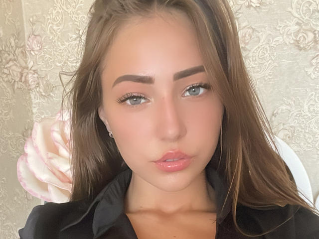 Miliaana has shiny brown hair, thick eyebrows, blue eyes, glossy pink lips, and is wearing a black shirt. Her butt is not visible, only her face is shown.