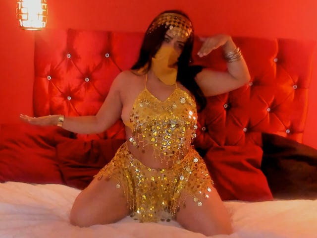 Anissa Ali, avec de grandes seins, est agenouillée sur un lit, vêtue d'une tenue brillante ornée de sequins dorés, portant un masque facial jaune et une pièce de tête, avec un dossier de lit rouge et capitonné en arrière-plan..