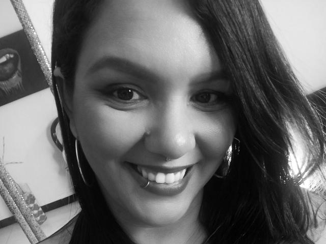 Black-and-white selfie of Capricornio69 with dark straight hair, hoop earrings, nose ring, and lip piercing, smiling at the camera. Capricornio69 has medium skin and visible teeth.