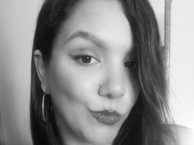 Black and white photo of Capricornio69 with dark, straight hair, hoop earrings, a nose piercing, and a lip piercing, making a pouty face with glossy lips.
