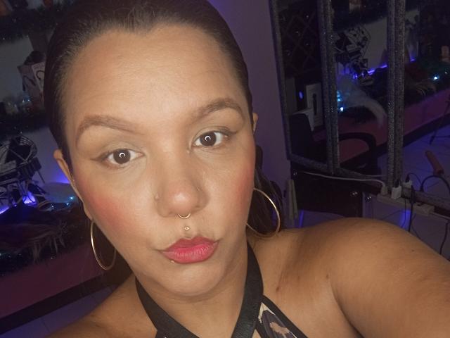 Capricornio69 with brown eyes, red lipstick, hoop earrings, nose piercing, and black lace halter top, showing a serious expression, in a dimly lit room.