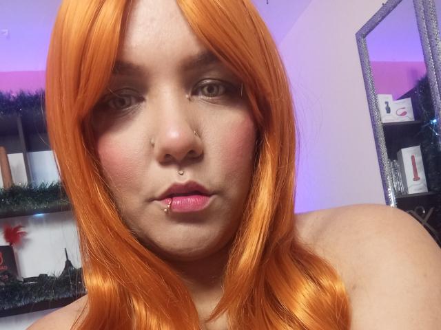 Capricornio69 with orange hair, nose ring, lip piercing, and pink lipstick, showing a serious expression. Visible bare shoulders. Reflective mirror in the background.
