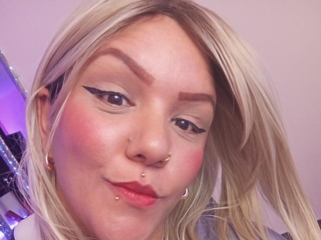 Capricornio69 has blonde hair, bold black eyeliner, pink blush, nose piercings, lip piercings, and gold earrings, with a slight smirk and a cute, attractive face.