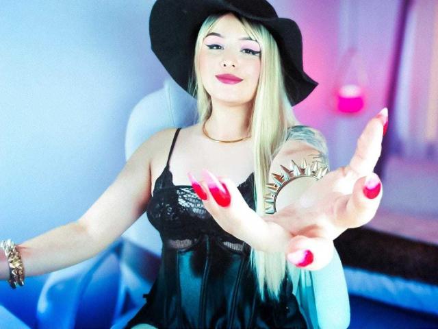 SophieLindemann with long blonde hair, black hat, black lace dress, red nails, and sun tattoo on right arm, pointing forward, smiling with pink lipstick.