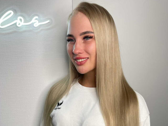 RamonaXKitty a de longs cheveux blonds, des yeux bleus et un sourire mignon. Elle porte une chemise blanche avec un logo noir, qui révèle ses petites seins. Elle se tient près d'un panneau lumineux..