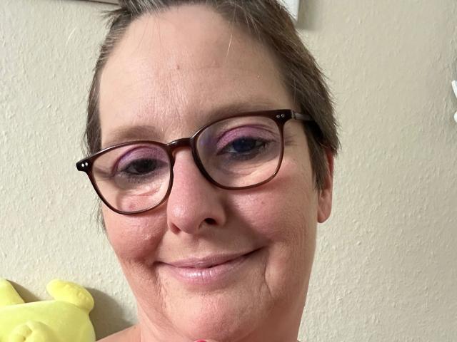 Chantallsweat-hot with glasses, purple eyeshadow, and a slight smile, showing wrinkles on her face and a yellow plush toy in the bottom left corner.