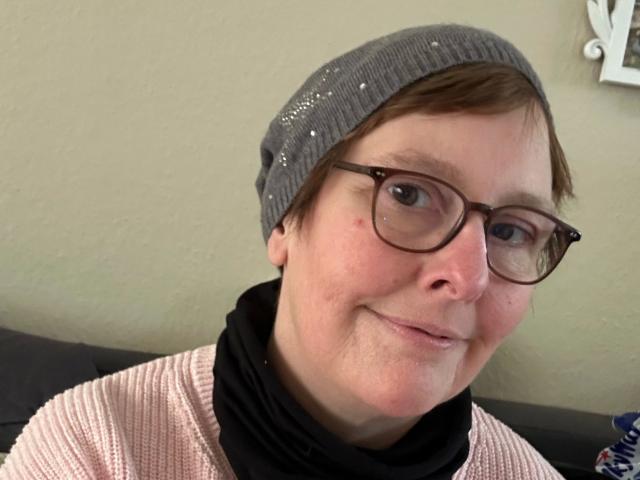 Chantallsweat-hot has short brown hair, wears a gray beanie, black glasses, and a pink knit sweater. Smiling with a slight nose freckle. Beige wall in background.