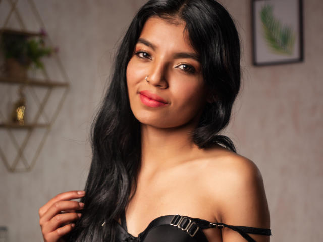 Emily Johanas mit langem, schwarzem Haar, braunen Augen und pinkem Lippenstift, trägt ein schwarzes Top mit einem Schulterauschnitt, lächelt sanft und hält ihr Haar. Brüste sind teilweise sichtbar. Warmes, indirektes Licht im Innenbereich..