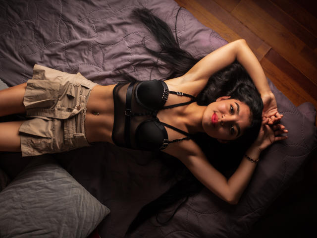 Emily Johanas est allongée sur un lit, les bras au-dessus de la tête, portant un soutien-gorge noir et un short beige, avec de longs cheveux noirs, du rouge à lèvres, un sol en bois brun, un oreiller gris et une couette foncée..