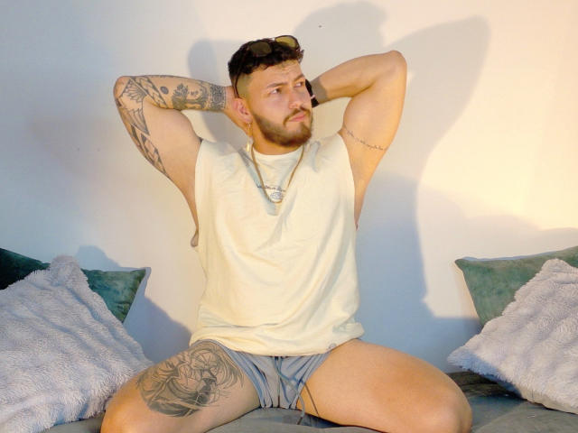 JaxRagnar, a tattooed dude with a beard, sits on a couch, arms behind his head, wearing a white tank and gray shorts, legs spread, looking slightly annoyed.