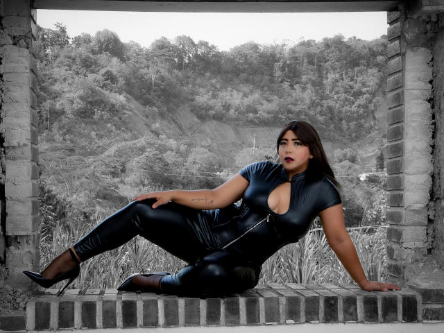 SamyWalker is laying in a brick frame wearing a tight black latex outfit with a zipper, showing her tits through a cutout, in black heels, looking sexy.