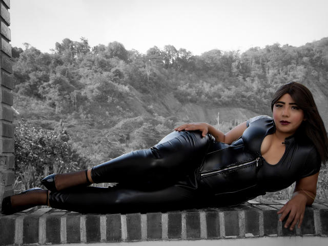 SamyWalker is laying on a brick ledge in black leather pants and a zipper-front top, exposing her tits. She has dark hair, black heels, and a serious expression.