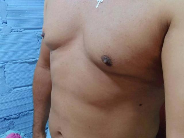 LoganModel's bare, tan-skinned chest with small, dark nipples and a subtle belly button. A silver cross necklace rests on his upper chest. His right arm is partially visible.
