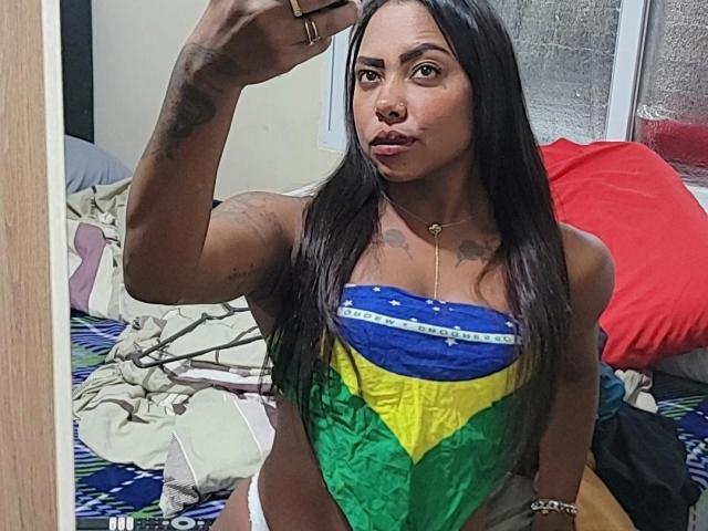 KarenEbony with long black hair, wearing a Brazilian flag strapless top, taking a selfie. She has tattoos on her arm and chest, and is pouting with her lips.