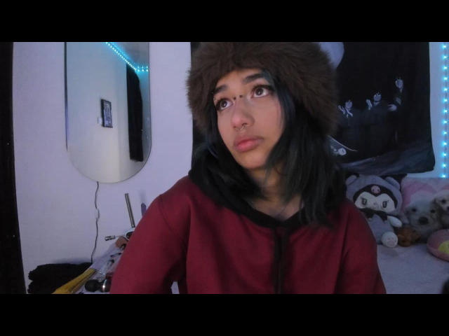 IsisJonnes69 with dark hair, wearing a brown furry hat and red shirt, has brown eyes and a nose ring, standing in a room with stuffed animals.