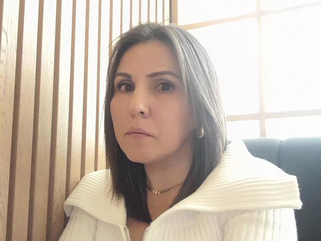 AdelaShery has medium-length black hair, brown eyes, and a neutral expression. She's wearing a white ribbed hoodie with a zipper and a gold necklace.