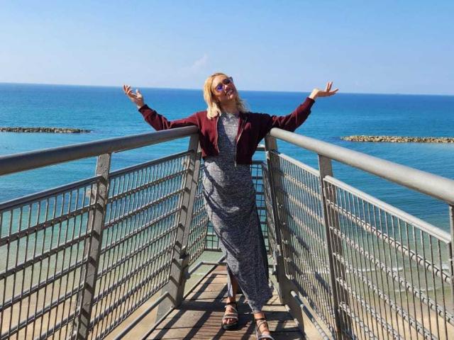 FreankyQueen stands on a metal bridge, arms out, wearing a gray dress, red jacket, and sunglasses, with a blue ocean and clear sky in the background.