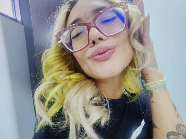 FreankyQueen, con cabello rubio y amarillo, usando gafas rojas y un piercing en la nariz, sonriendo con los ojos cerrados, camisa negra y tatuajes en el brazo derecho..