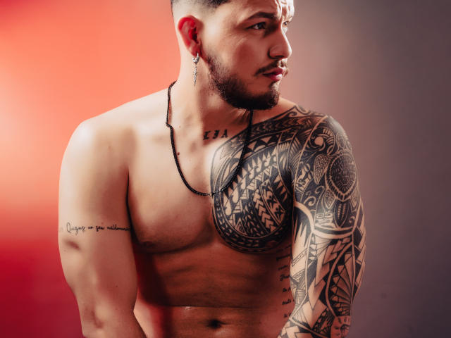 JaxRagnar has a muscular, tattooed chest and arm, short dark hair, and a beard. He's shirtless with a black necklace and a side profile. His arm tattoos are detailed.