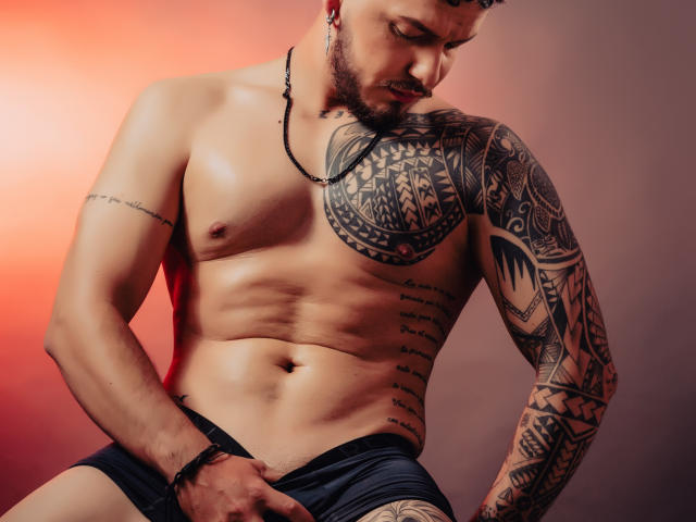 JaxRagnar, muscular, tattooed, shirtless, with black shorts, pulling down his shorts to reveal his thick, uncut cock. Red and orange lighting. His right hand grips his cock.