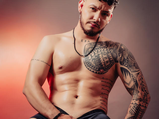 JaxRagnar, shirtless, muscular, tattooed, brown hair, beard, necklace, slight smirk, right hand on black pants, left arm tattoos, right arm tattoo, tan skin, red light background.