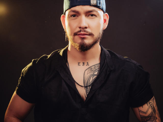 JaxRagnar has a black shirt, tattoos on chest and arm, and a beard, wearing a blue cap. His chest tattoo reads "13A" and he has a necklace.