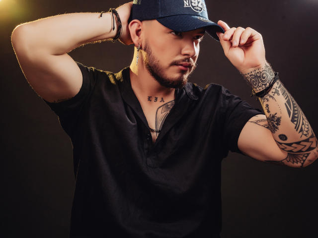JaxRagnar, a tattooed dude, wears a black shirt and cap, adjusting his cap with hands up, showing off arm tattoos, and sporting a beard. He's looking serious and hot.