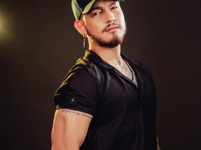 JaxRagnar with a green cap, black shirt, tattoo on right arm, and gold hoop earring, looking down with a slight smirk, brown background.