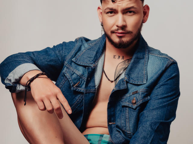 JaxRagnar, shirtless with a blue denim jacket, has tattoos on his chest and neck, a beard, and is wearing green underwear, sitting with a relaxed, confident expression.