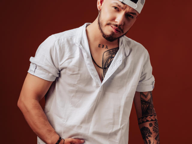 JaxRagnar with a white shirt, tattooed arms, and a backward cap, has a hand on his hip and a playful smirk. Chest tattoo, short beard, brown background.