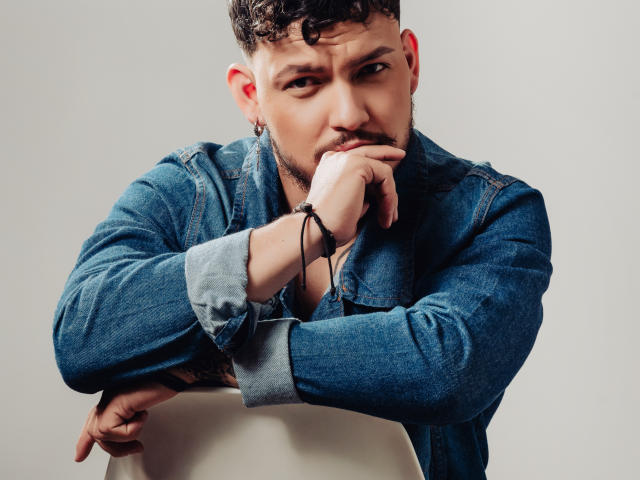 JaxRagnar, with curly brown hair and a beard, wearing a blue denim shirt, rests his chin on his hand, looking serious, sitting on a white chair.